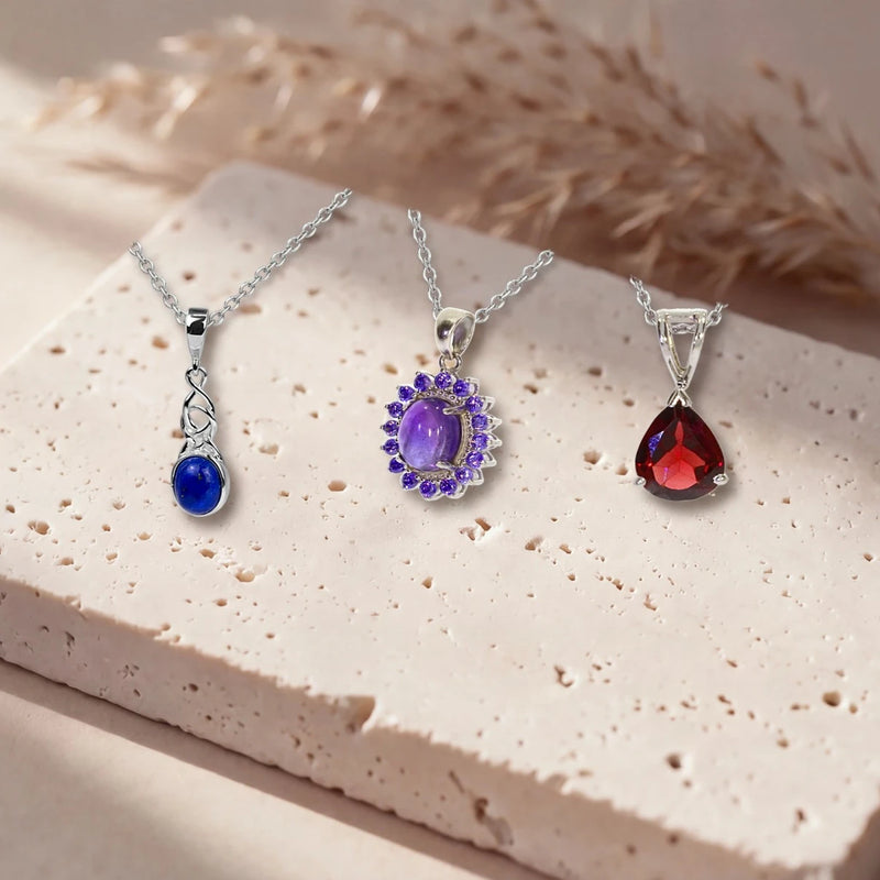 Pendentifs argent et pierre naturelle en lapis-lazuli, améthyste et grenat sur leur chaîne, posés sur une dalle de travertin