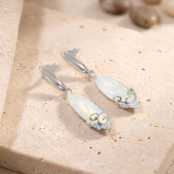 Boucles d'oreilles en pierre de lune cabochon navette sur argent 925 rhodié, paire posée sur une dalle de travertin