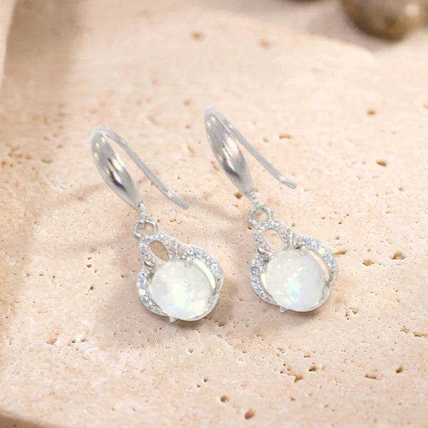 Boucles d'oreilles pendantes en pierre de lune ovale sur crochet en argent 925 rhodié, paire posée sur une dalle de travertin