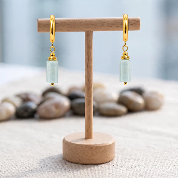 Boucles d'oreilles pierre bleu clair, tube d'aigue-marine sur créole dorée, présentées sur un support en bois