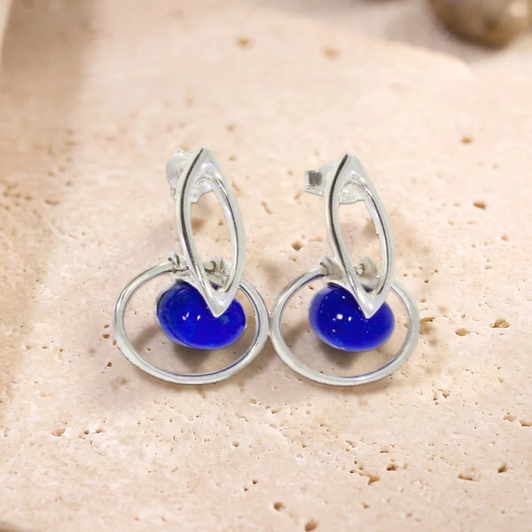 Boucles d'oreilles boule de lapis-lazuli dans un cercle en argent 925 rhodié, paire posée sur une dalle de travertin