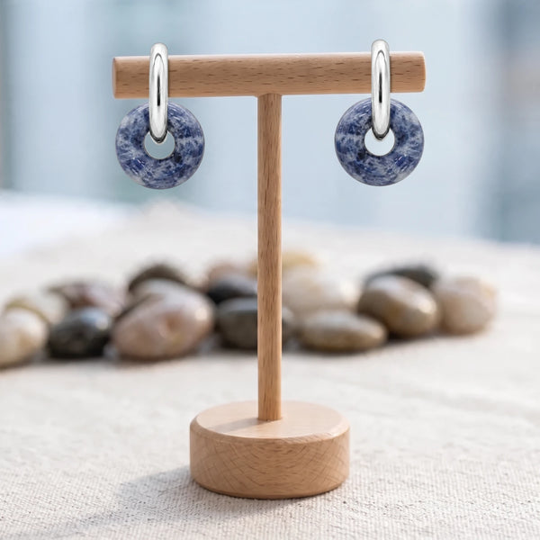 Boucles d'oreilles pierre bleue, donut de sodalite 15 mm sur créole argentée, présentées sur un support en bois