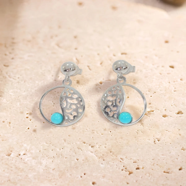 Boucles d'oreilles en turquoise sur cercle ajouré en argent 925 rhodié, paire posée à plat sur une dalle de travertin