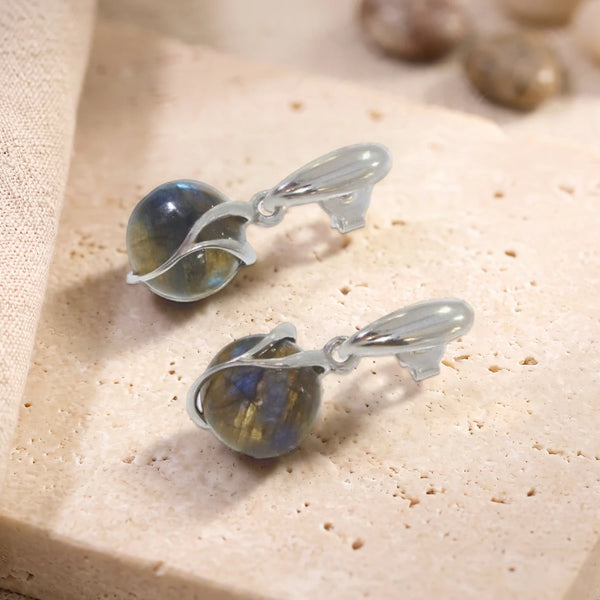 Boucles d'oreilles boule de labradorite en argent 925 rhodié, paire posée sur une dalle de travertin