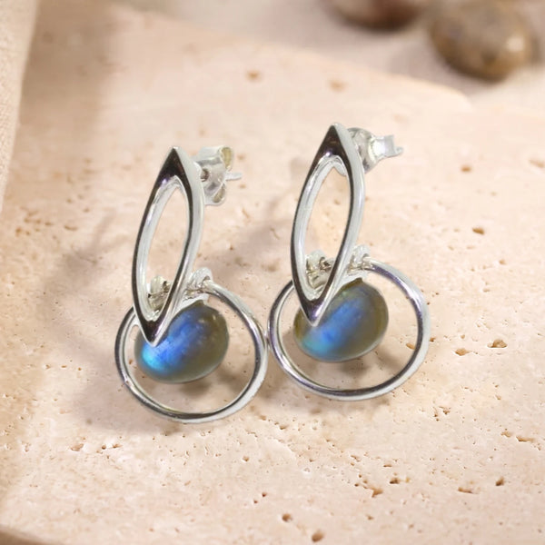 Boucles d'oreilles boule de labradorite dans un cercle en argent 925 rhodié, paire posée sur une dalle de travertin