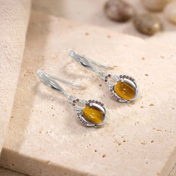 Boucles d'oreilles pendantes en œil de tigre ovale sur crochet en argent 925 rhodié, paire posée sur une dalle de travertin