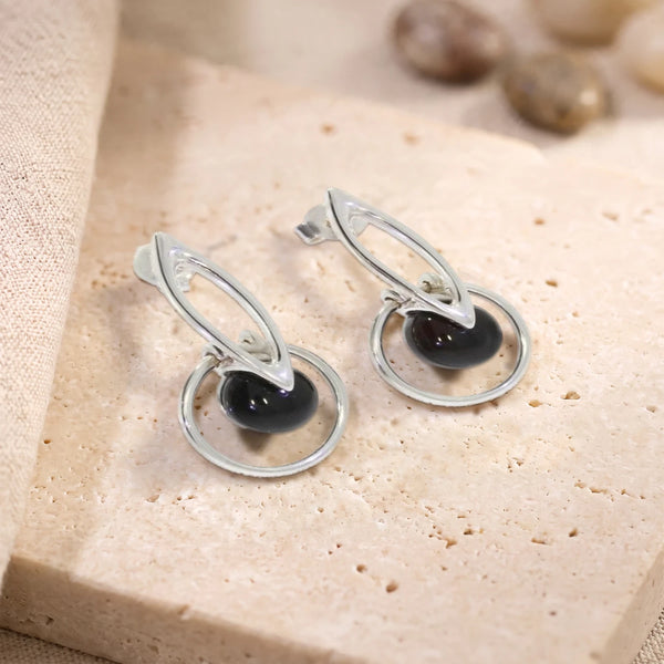 Boucles d'oreilles boule d'onyx noir dans un cercle en argent 925 rhodié, paire posée sur une dalle de travertin