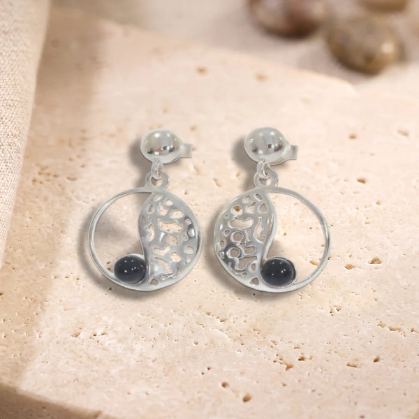 Boucles d'oreilles en onyx noir sur cercle ajouré en argent 925 rhodié, paire posée à plat sur une dalle de travertin