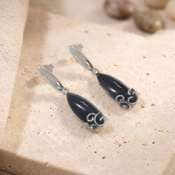 Boucles d'oreilles en onyx noir cabochon navette sur argent 925 rhodié, paire posée sur une dalle de travertin