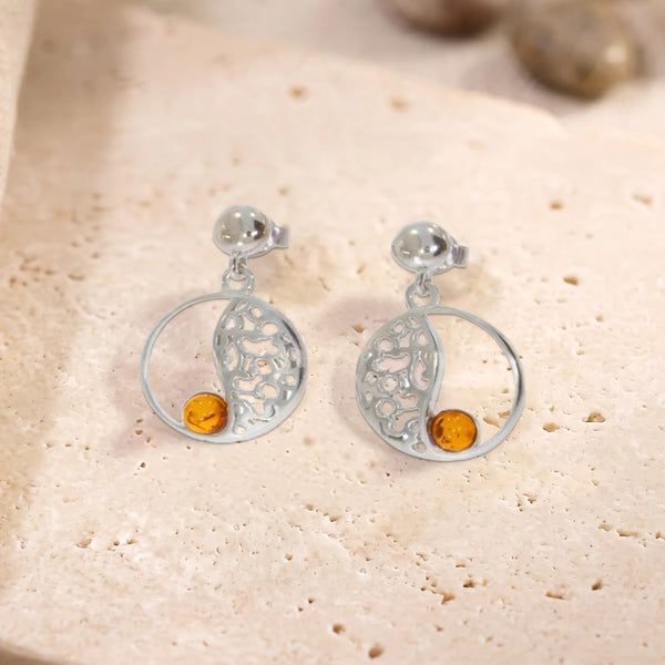 Boucles d'oreilles en ambre sur cercle ajouré en argent 925 rhodié, paire posée à plat sur une dalle de travertin