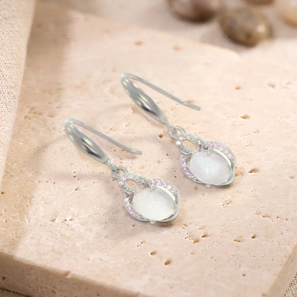 Boucles d'oreilles pendantes en quartz rose ovale sur crochet en argent 925 rhodié, paire posée sur une dalle de travertin
