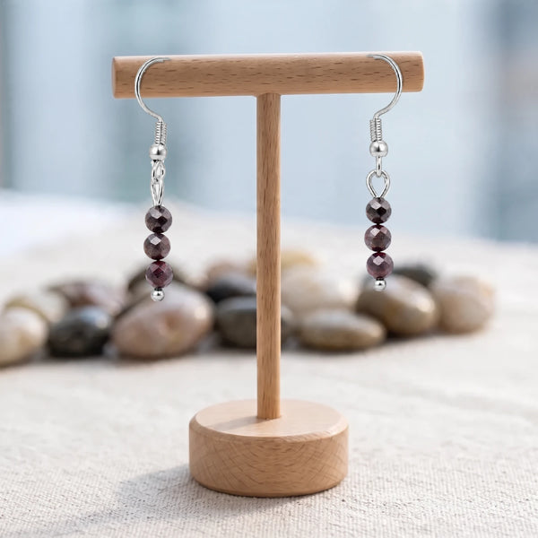 Boucles d'oreilles pierre rouge, trois perles facettées de grenat sur crochet argenté, présentées sur un support en bois
