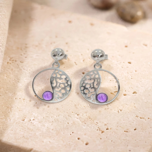 Boucles d'oreilles en améthyste sur cercle ajouré en argent 925 rhodié, paire posée à plat sur une dalle de travertin