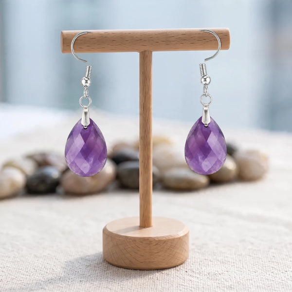 Boucles d'oreilles pierre violette, goutte d'améthyste facettée 18 mm sur crochet argenté, présentées sur un support en bois