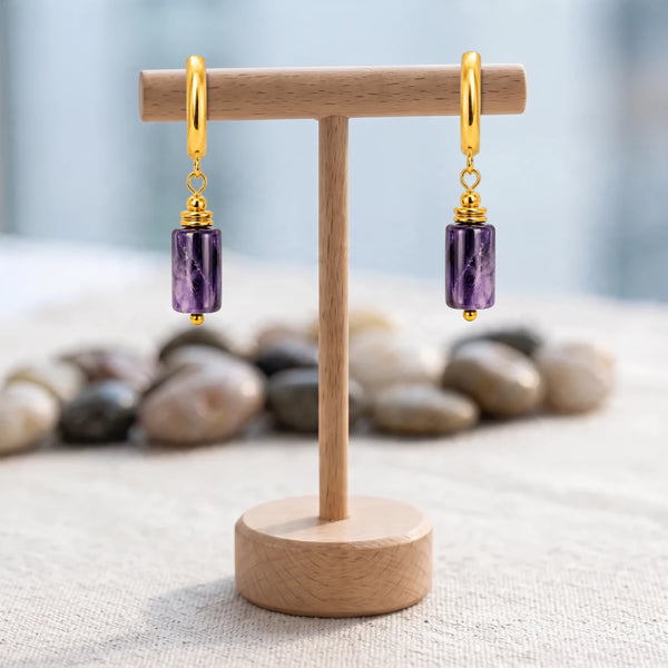 Boucles d'oreilles pierre violette, tube d'améthyste sur créole acier doré, présentées sur un support en bois
