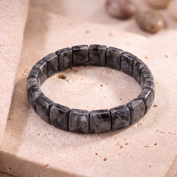 Bracelet larvikite homme en plaques de 14 mm, pierre grise aux reflets bleus, posé sur une dalle de travertin
