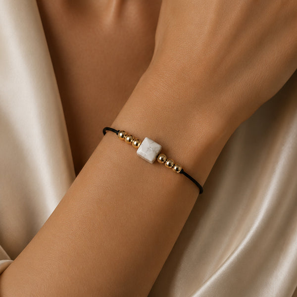 Bracelet pierre naturelle howlite blanche porté au poignet d'une femme, cube blanc veiné de gris et cordon doré CUBE®
