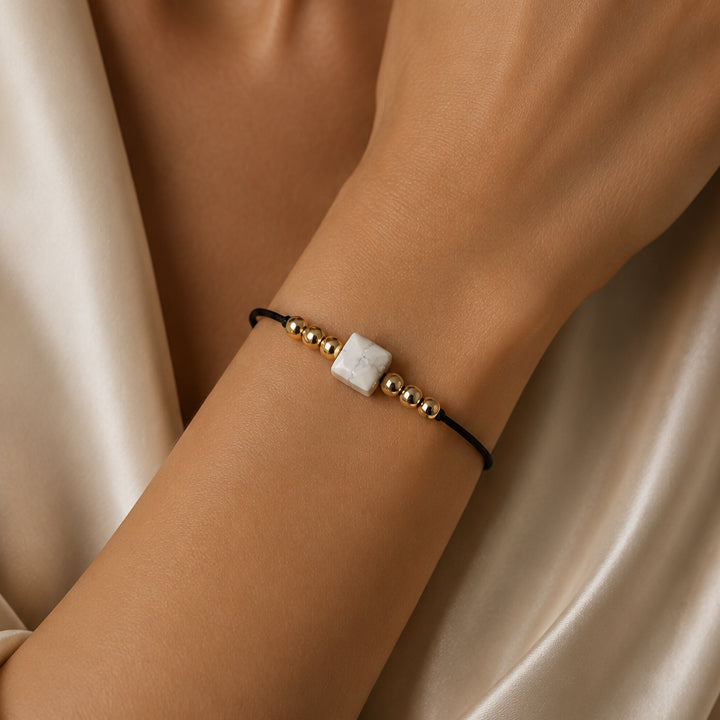 Bracelet pierre naturelle howlite blanche porté au poignet d'une femme, cube blanc veiné de gris et cordon doré CUBE®