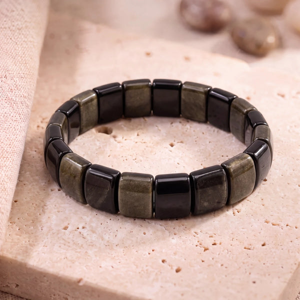 Bracelet obsidienne dorée en plaques de 14 mm, pierre noire au reflet doré, posé sur du travertin