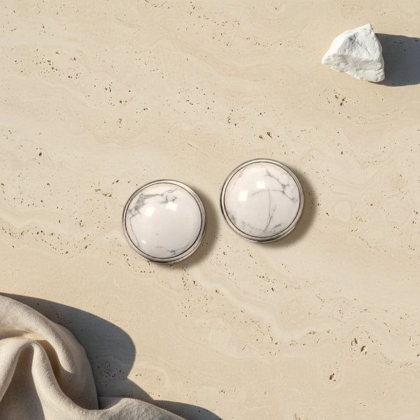 Cache bouton blanc en howlite veinée de gris, deux cabochons sertis d'acier posés à plat sur du travertin