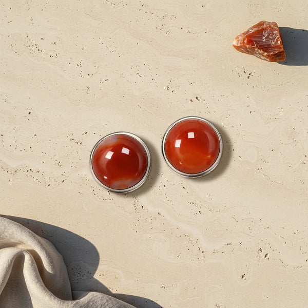 Cache bouton orange en cornaline naturelle, deux cabochons sertis d'acier posés à plat sur du travertin