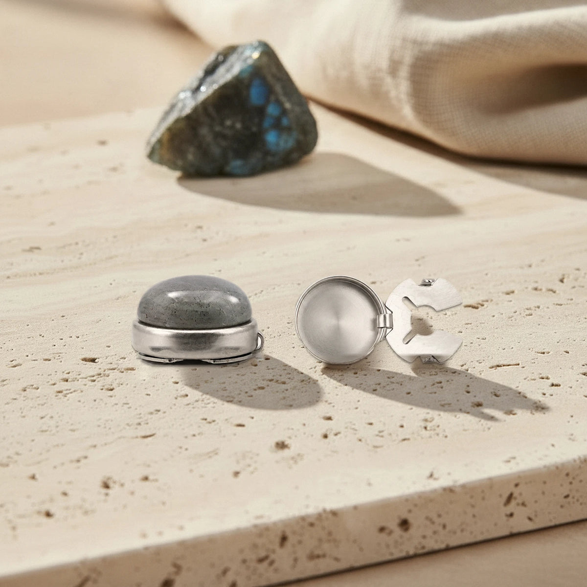 Couvre bouton en labradorite grise avec son fermoir à clip en acier ouvert, posé sur du travertin