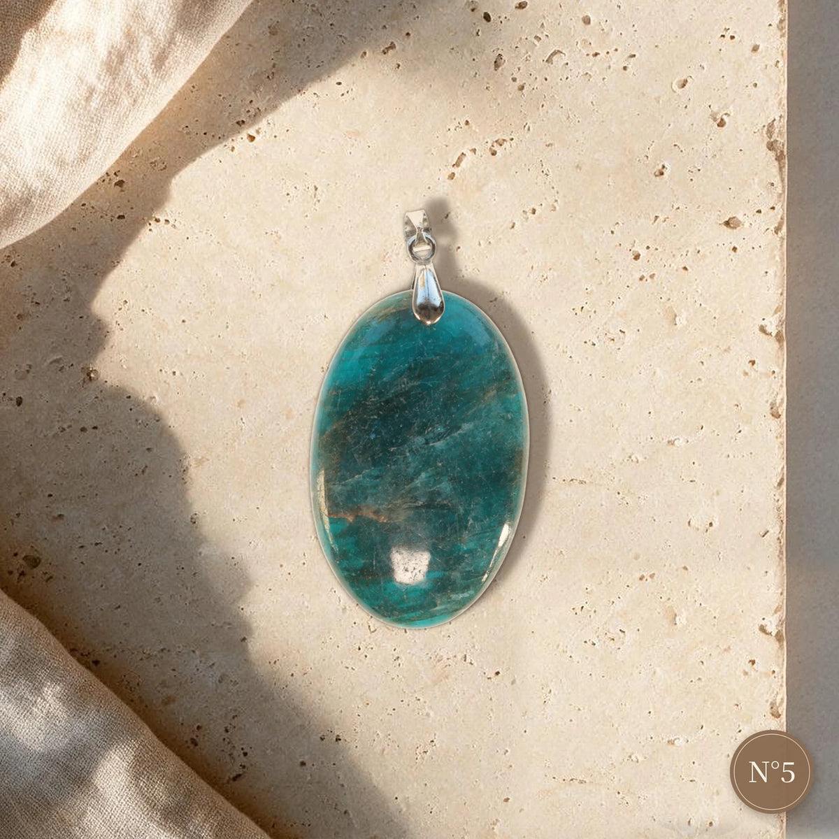 Pendentif apatite bleue pièce unique N°5, ovale bleu turquoise aux reflets sombres et bélière argentée sur travertin