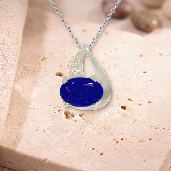 Pendentif lapis lazuli femme, modèle ovale facetté sur griffes, sur sa chaîne, posé sur une dalle de travertin