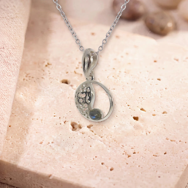 Petit pendentif labradorite, modèle médaillon ajouré vintage, sur sa chaîne, posé sur une dalle de travertin