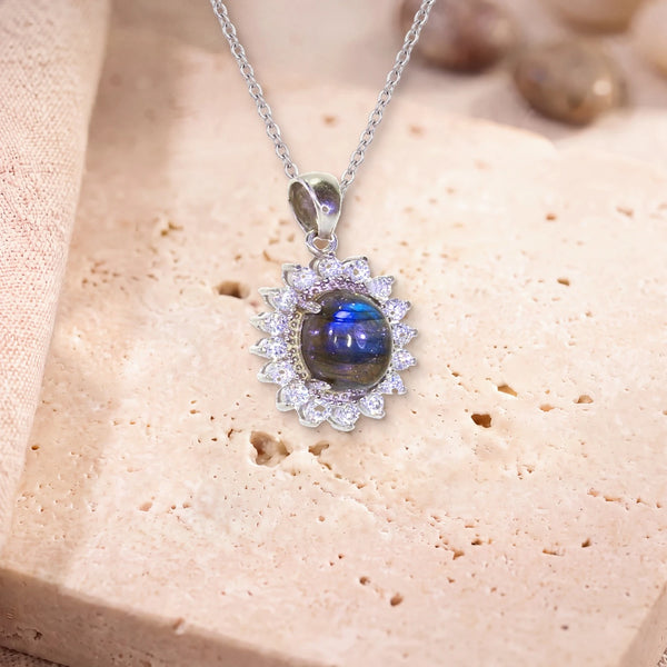Pendentif pierre labradorite, modèle médaillon ovale bordé de zircons, sur sa chaîne, posé sur une dalle de travertin