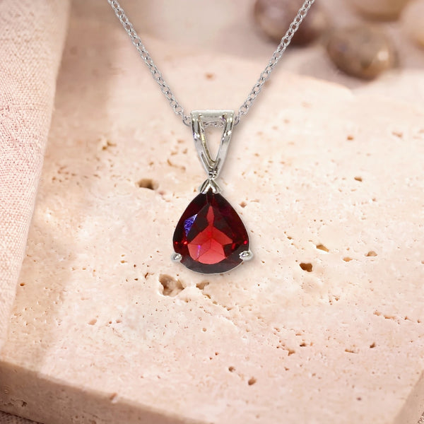 Pendentif grenat rouge, modèle petite goutte facettée, sur sa chaîne, posé sur une dalle de travertin
