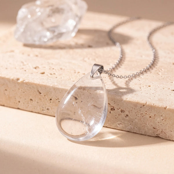 Pendentif pierre transparente en cristal de roche, goutte plate polie sur chaîne argentée, posé sur travertin