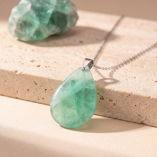 Pendentif pierre verte en fluorine, goutte plate polie sur chaîne argentée, posée sur une dalle de travertin