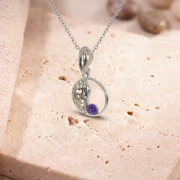 Collier pendentif améthyste, modèle médaillon ajouré vintage, sur sa chaîne, posé sur une dalle de travertin