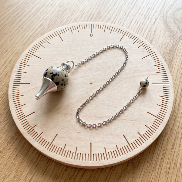 Pendule blanc séphoroton en jaspe dalmatien posé sur une planche de radiesthésie en bois