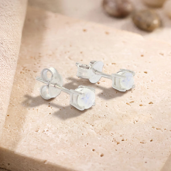 Puces d'oreilles en pierre de lune ronde 4 mm sur argent 925 rhodié, paire posée sur une dalle de travertin