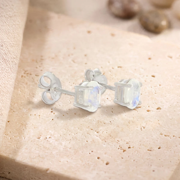 Puces d'oreilles en pierre de lune ovale 7 x 5 mm sur argent 925 rhodié, paire posée sur une dalle de travertin