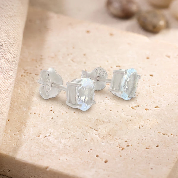 Puces d'oreilles en aigue-marine ovale 7 x 5 mm sur argent 925 rhodié, paire posée sur une dalle de travertin