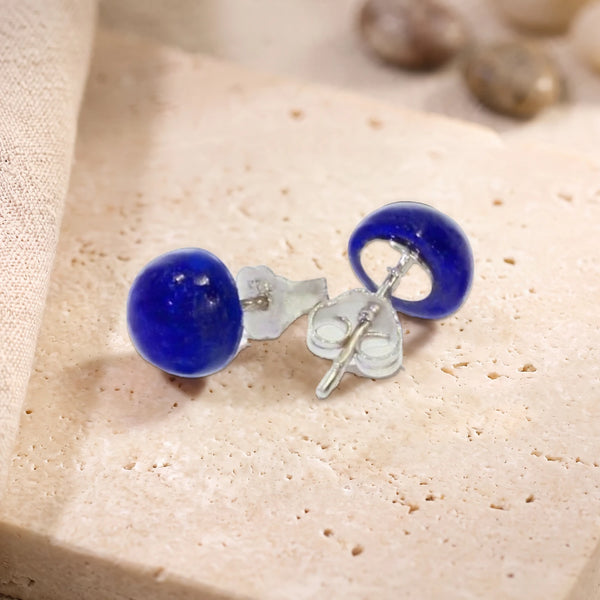 Puces d'oreilles en lapis-lazuli bleu, cabochon rond 8 mm sur argent 925 rhodié, paire posée sur une dalle de travertin