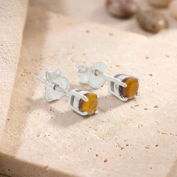 Puces d'oreilles en œil de tigre rond 4 mm sur argent 925 rhodié, paire posée sur une dalle de travertin
