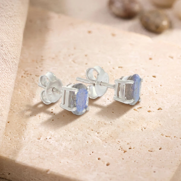 Puces d'oreilles en labradorite ovale 7 x 5 mm sur argent 925 rhodié, paire posée sur une dalle de travertin